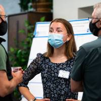 Scholar presenting poster to two guests, all wearing masks.